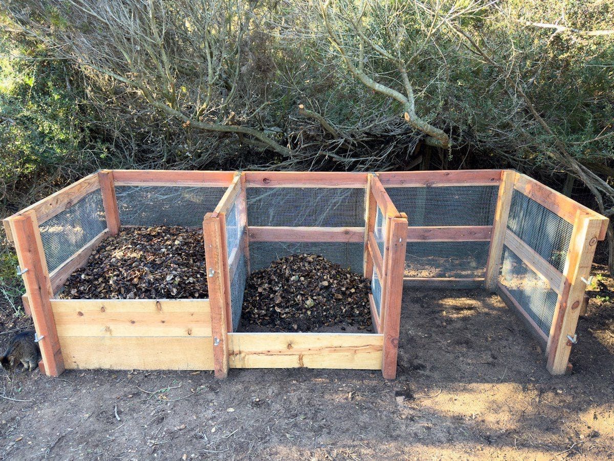 Three-bin wooden compost system with mesh sides, filled with decomposing organic matter, outdoors.