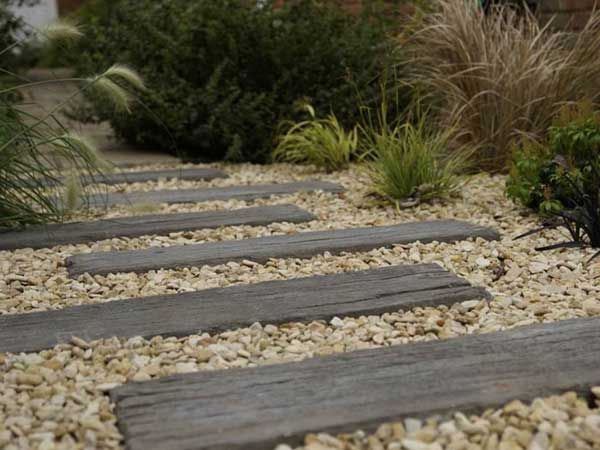 Wooden planks form a path through a gravel bed, surrounded by ornamental grasses and greenery.