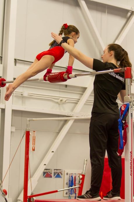 Une gymnaste aux barres asymétriques, assistée par son entraîneur, vêtue de rouge et de noir, en intérieur.