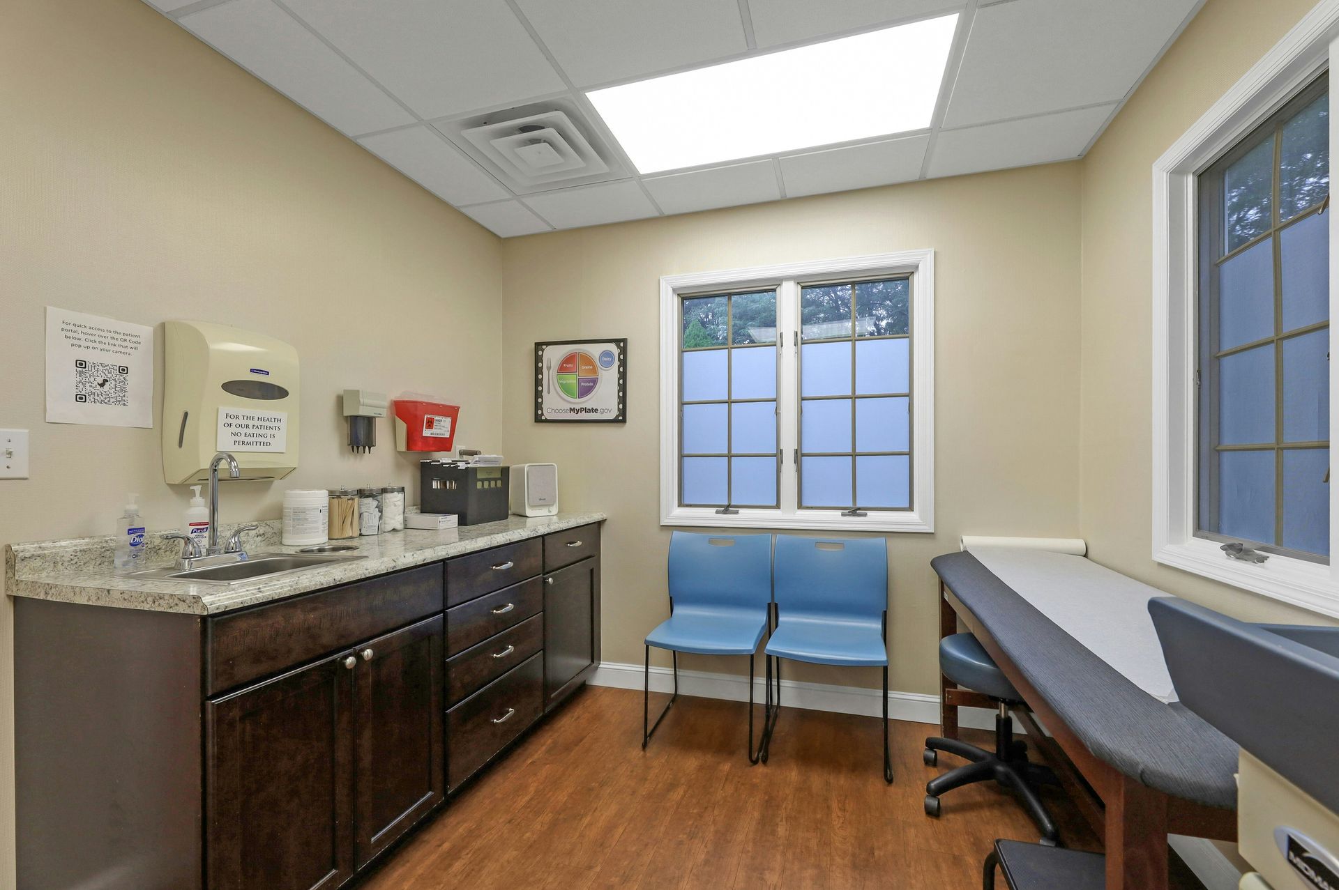 Exam room with brown cabinets, a sink, and an examination table. Two blue chairs face a window.