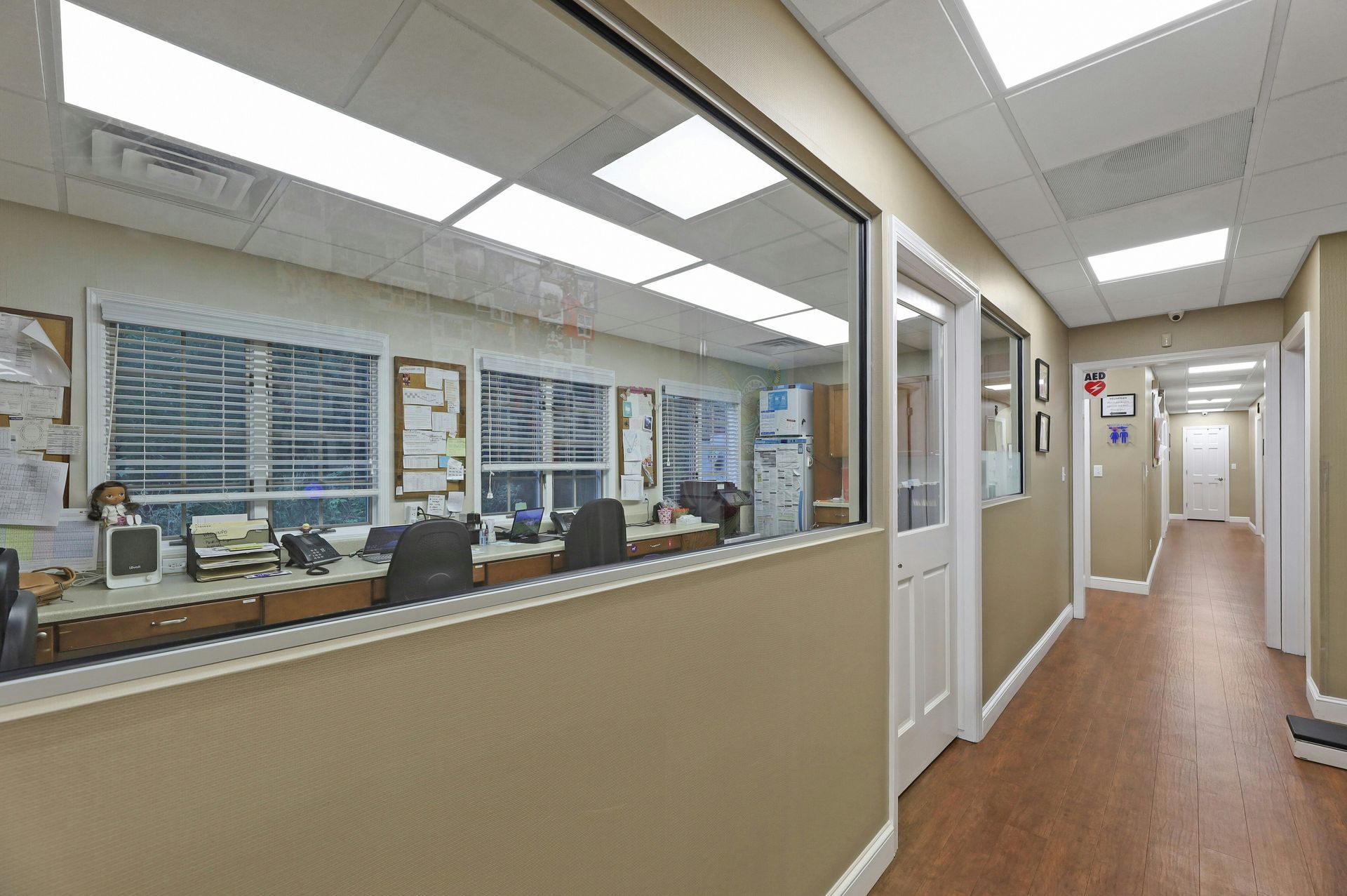 Hallway with office visible through a large window, white doors, brown wood-look floor, and ceiling lights.