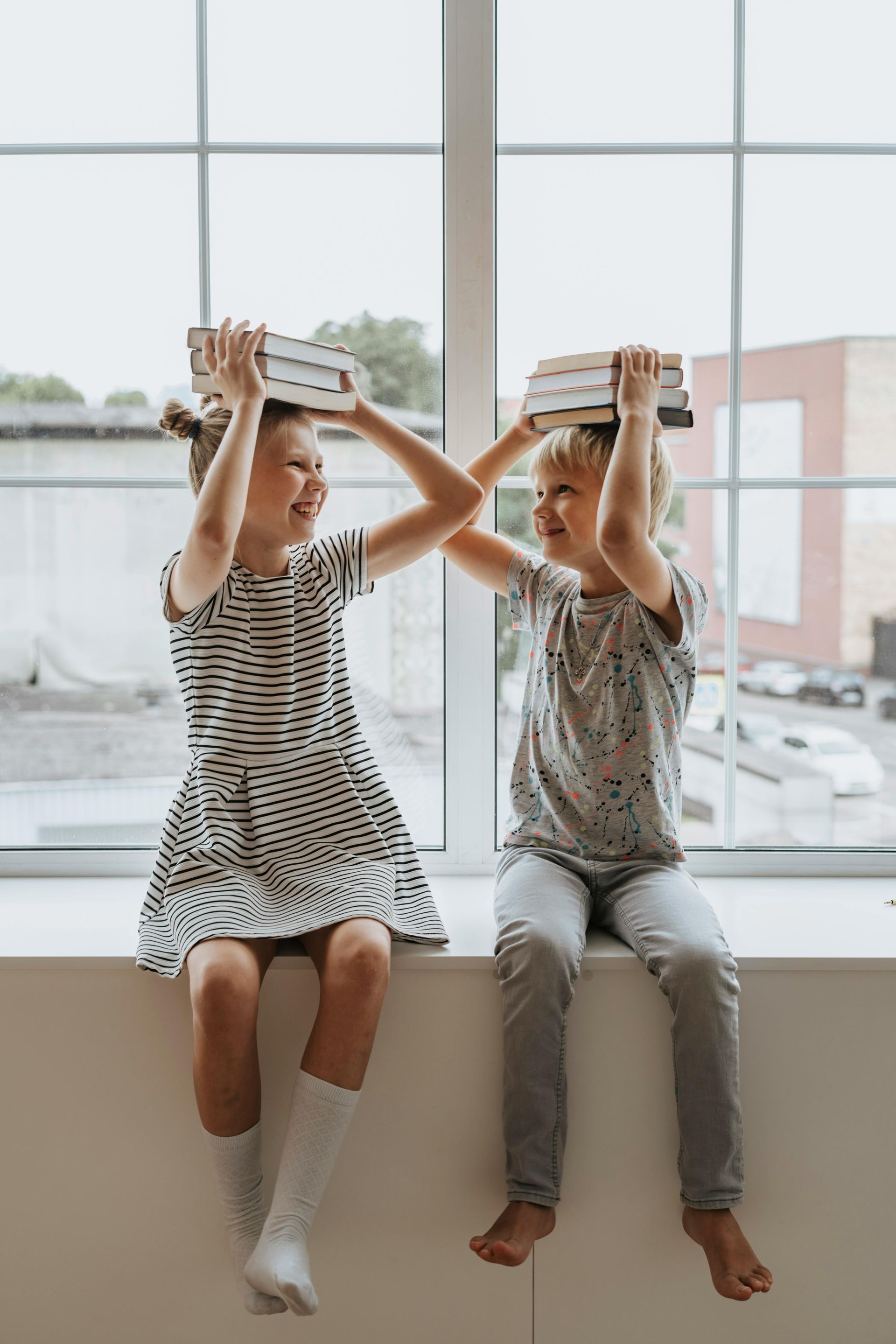 Two children sitting in front of a window with books on their heads.