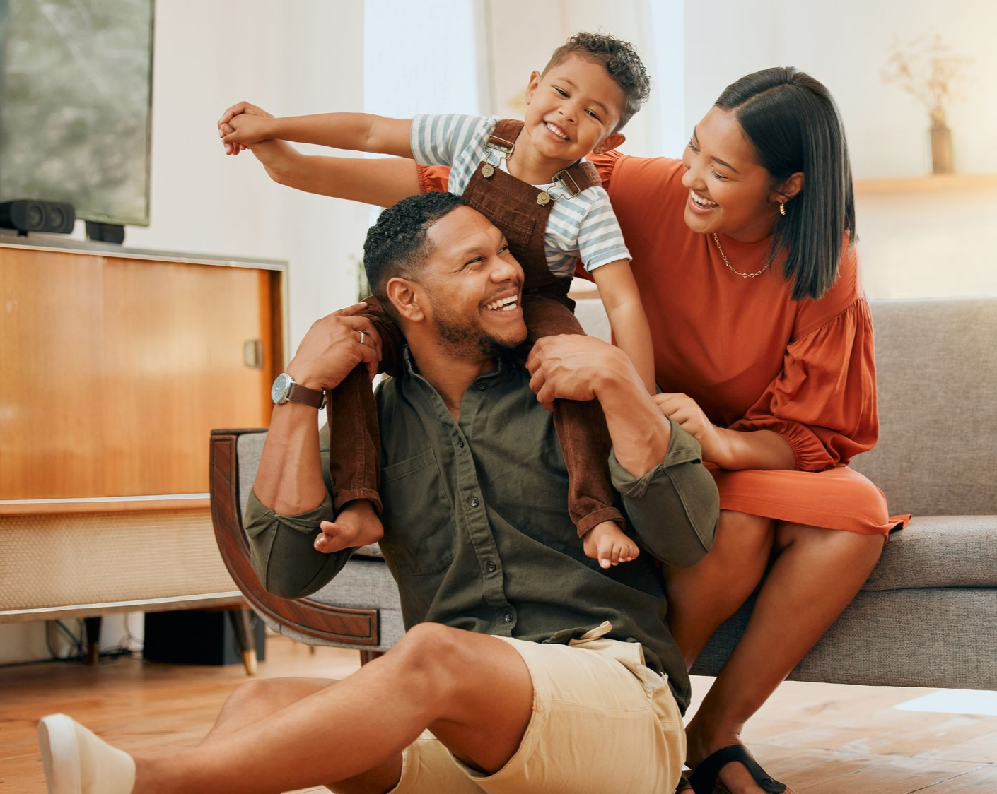 A man with a young boy on his shoulders is sitting on the floor. A woman is sitting behind them and they are all smiling.