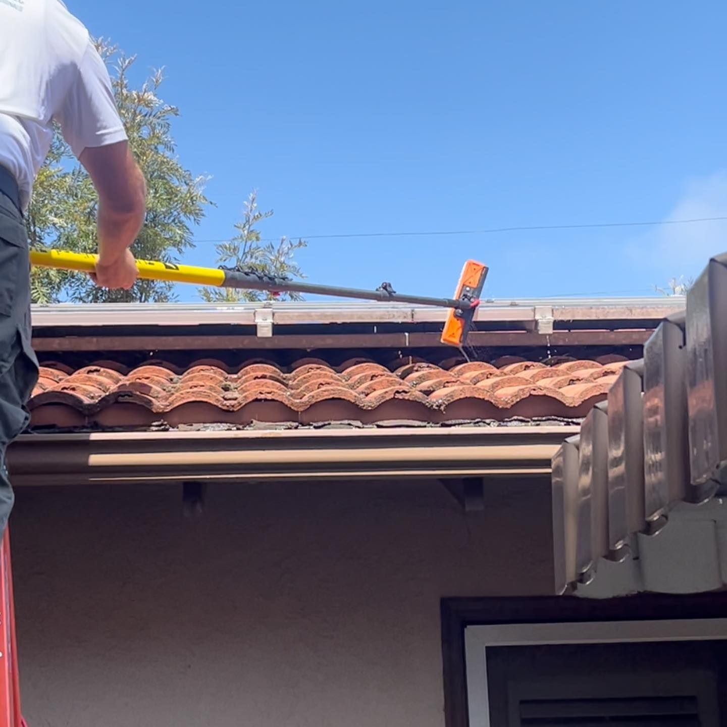 A man is standing on a ladder cleaning the roof of a house.