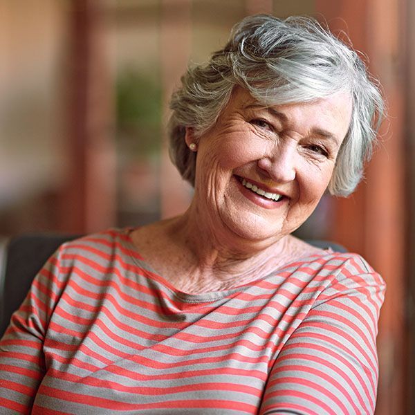 Smiling older woman in a coral-striped shirt, seated indoors against a blurred warm background