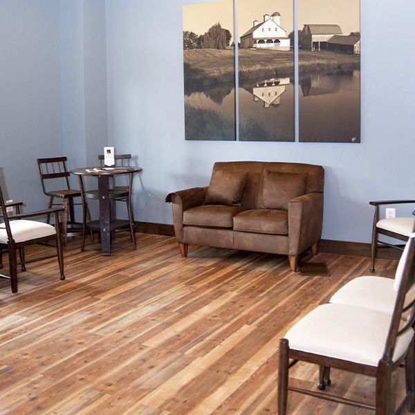 Empty waiting room with brown loveseat, wooden chairs, small table, and wall art on a wood floor