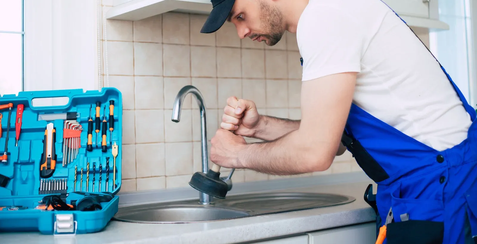 Plumber in blue overalls working on a kitchen faucet near a blue toolbox on a counter.