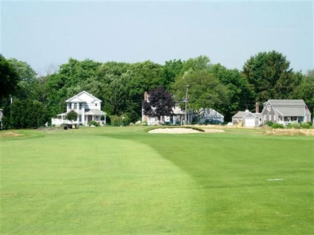 Wide green field with a few white houses and trees under a clear blue sky