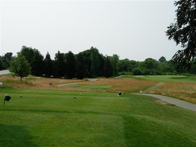 Grassy golf course fairway with sand traps, trees, and a paved cart path under a cloudy sky