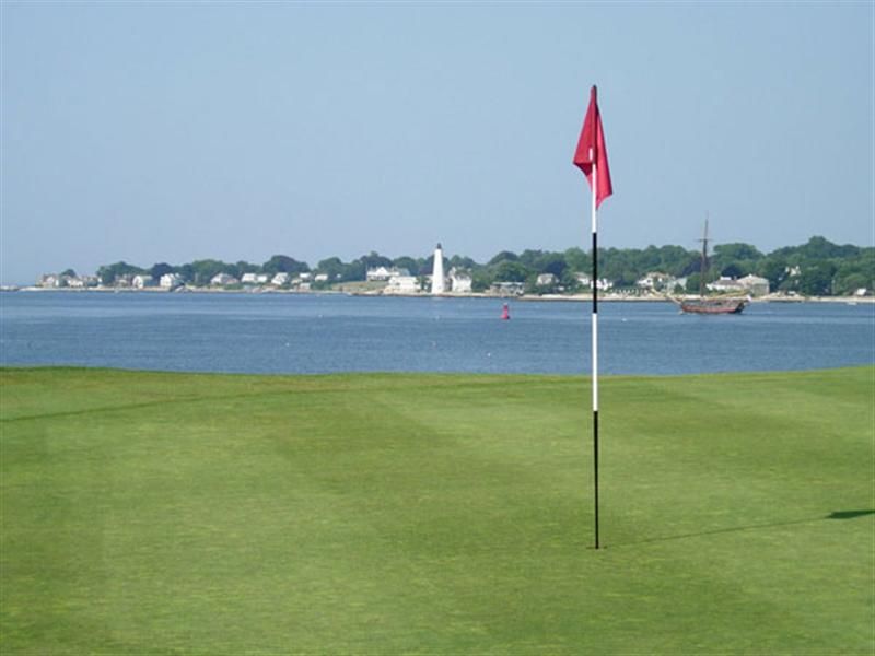 Golf green with a red flag by the water, shoreline and buildings in the distance
