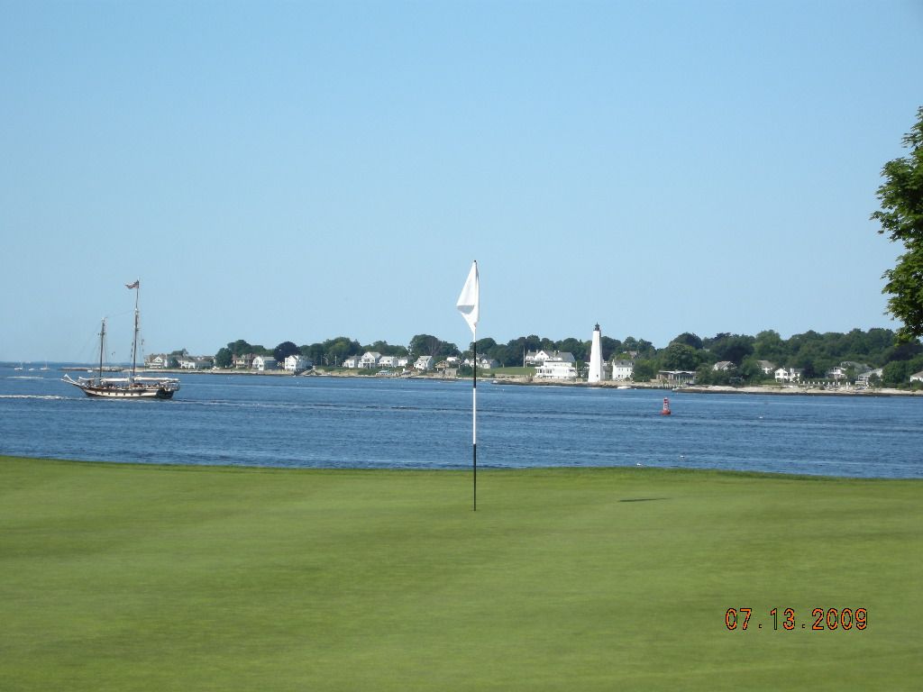 Grassy shoreline overlooking blue water with a sailboat and distant lighthouse under a clear sky.
