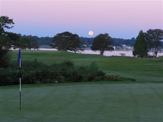 Moonrise over a golf course at dusk, with a flagstick and trees silhouetted against a pink sky