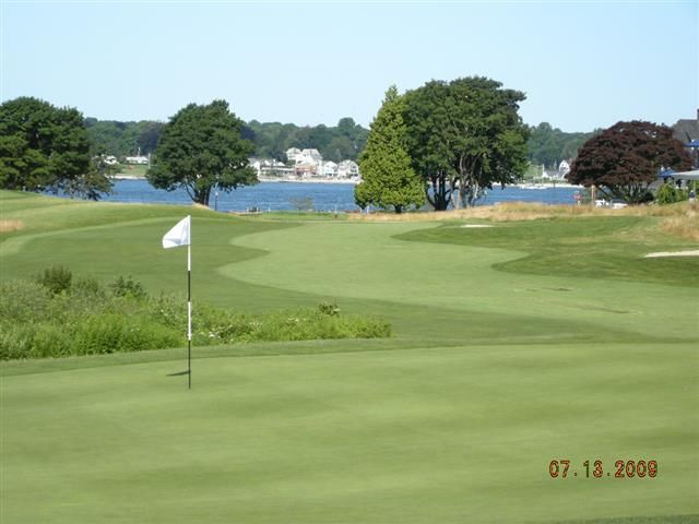 Golf green with flag by a lakeside, trees and houses in the background under a clear sky