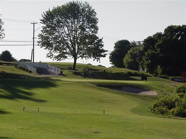 A green golf course with a sand bunker, trees, and a flag in the distance under a hazy sky