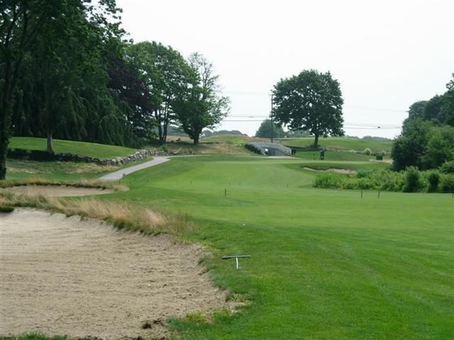 Golf course fairway with sand bunker, trees, and a paved cart path under cloudy skies