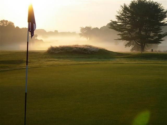 Golf course at sunrise with flagstick, green fairway, mist, and trees in the distance