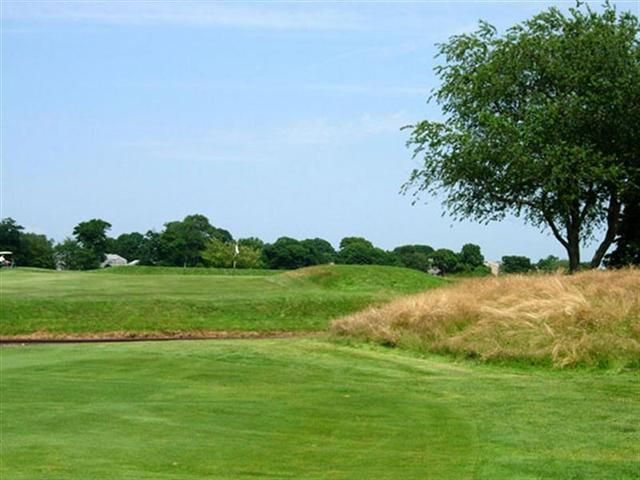 Lush green golf course with trees, a sand bunker, and a clear blue sky