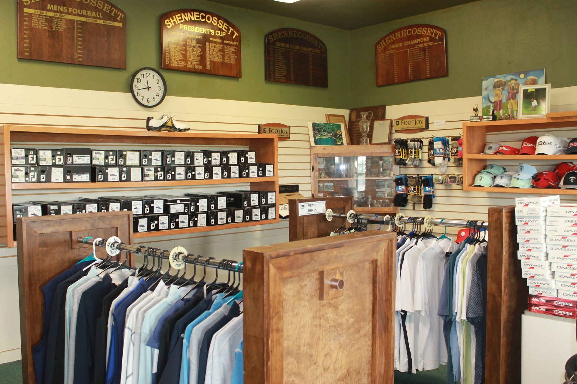 Small clothing store interior with racks of shirts, shelves of boxes, and framed signs on green walls.