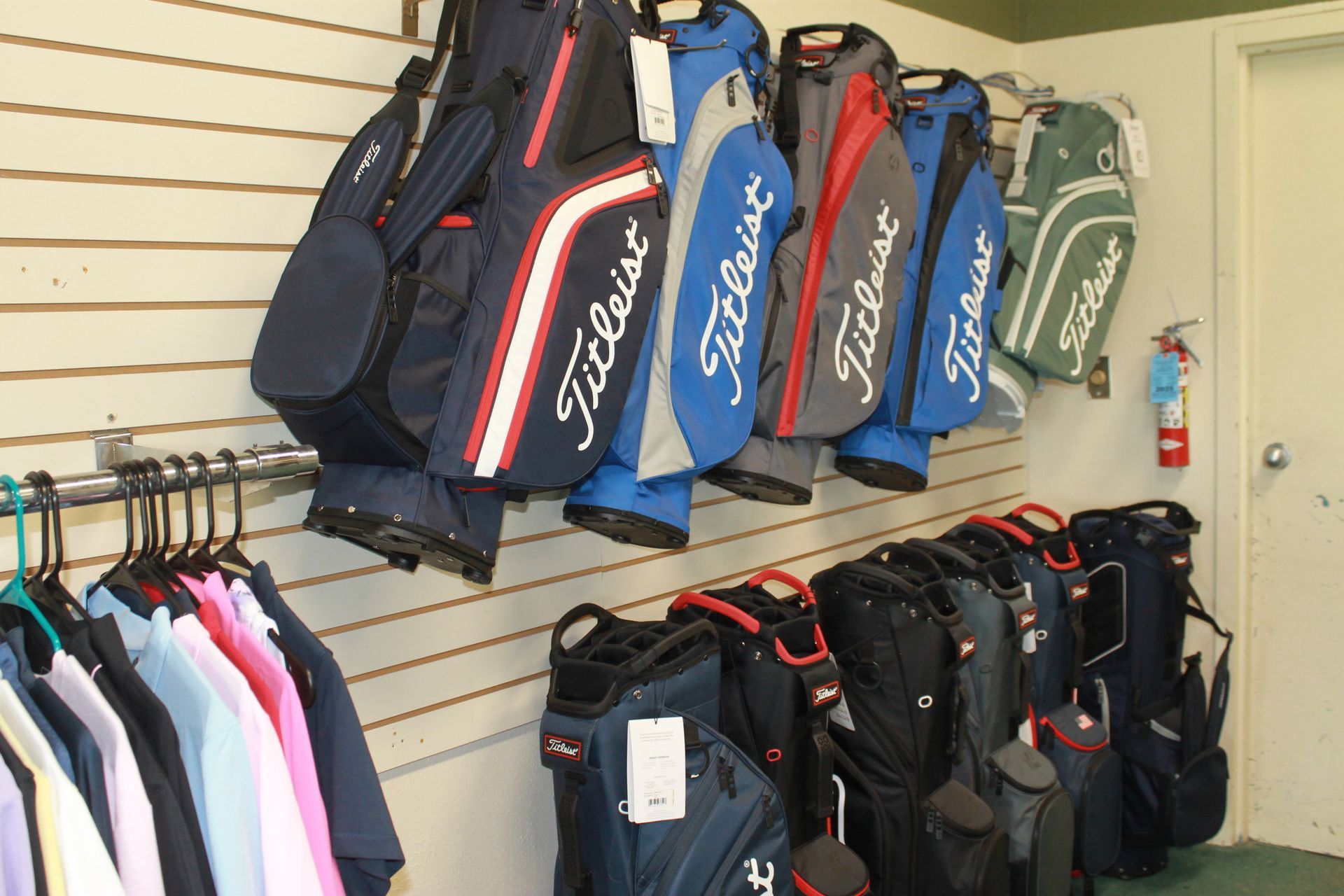 Golf bags hanging on wall racks in a sporting goods store, with clothing racks below.