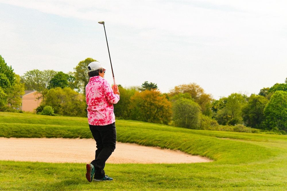 Golfer in pink shirt swinging a club near a sand bunker on a green course
