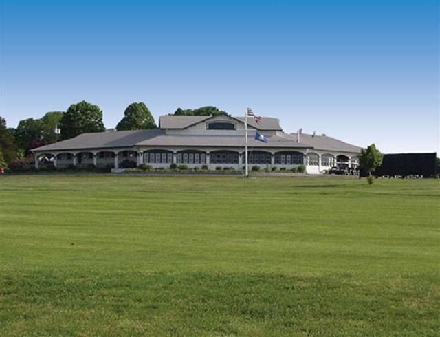 Large white clubhouse with a flagpole behind a wide green lawn under a clear blue sky