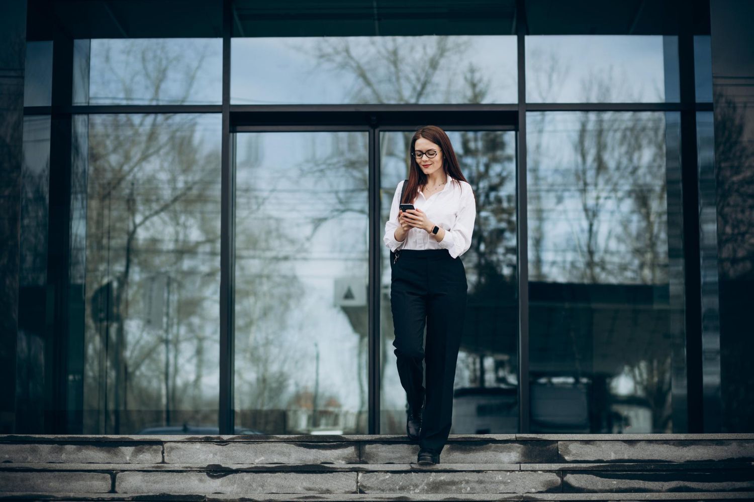Woman in a white shirt and black pants looking at her phone outside a glass building