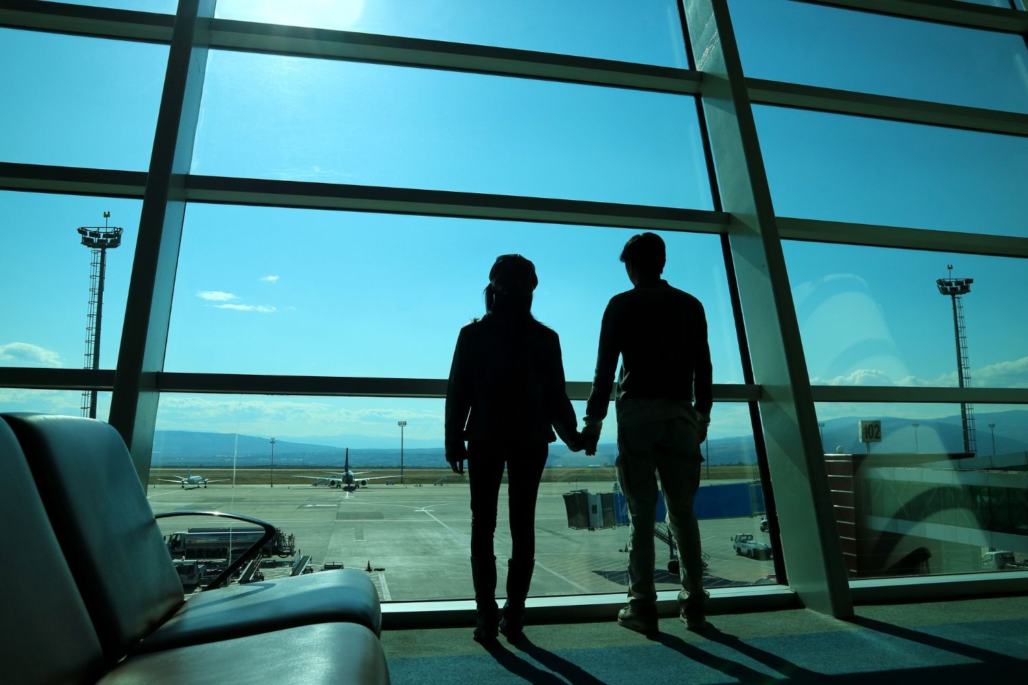 Two silhouetted travelers holding hands in an airport terminal, with blue sky and tarmac beyond the windows