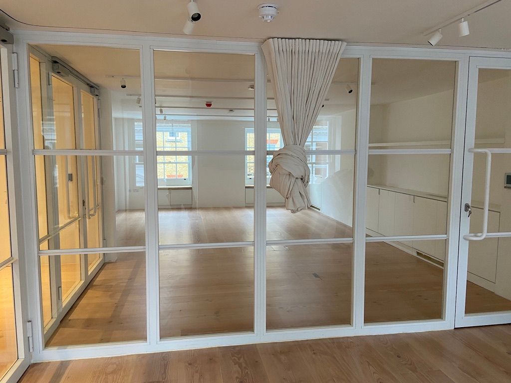 Empty modern room with white-framed glass partition and wood floors, bright windows in back.