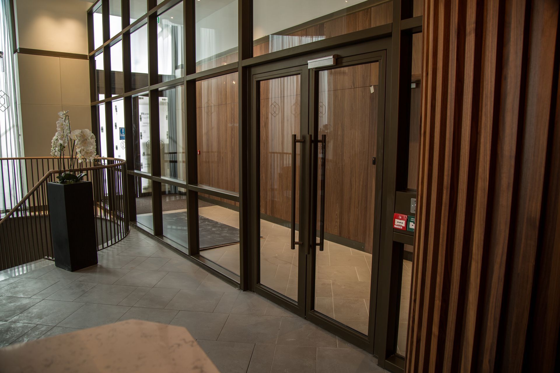 Glass entrance doors with metal frames in a modern lobby, beside vertical wooden slats.