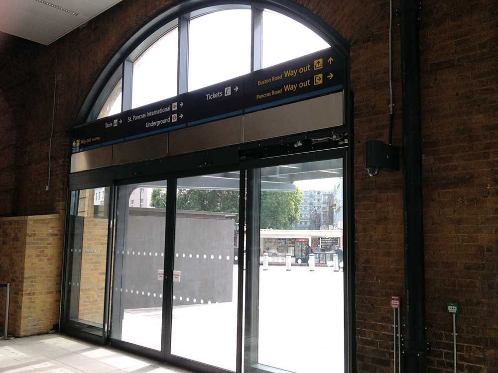 Glass station entrance with arched window and brick walls, doors open to a bright outdoor platform area