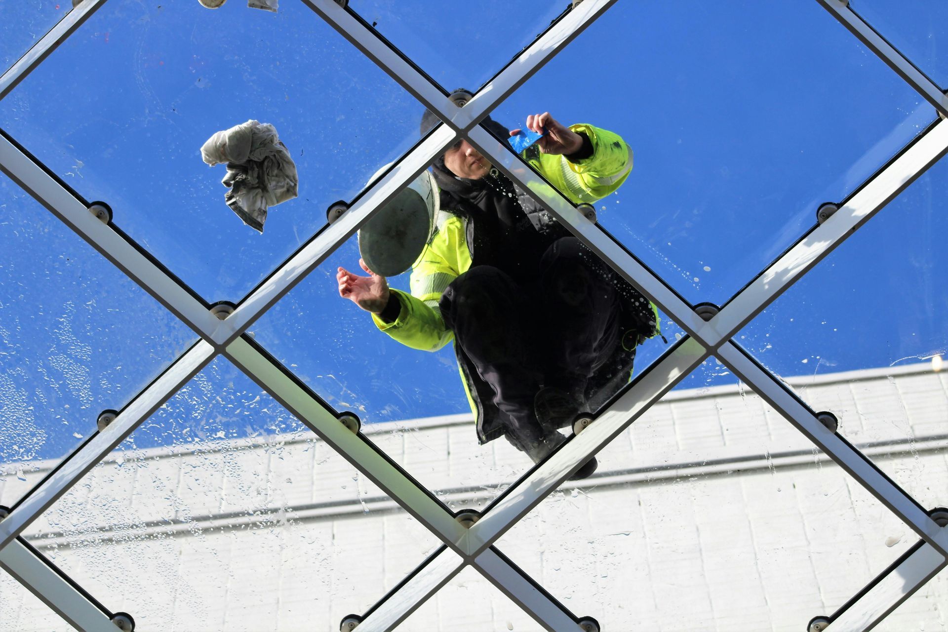 Person in a neon jacket cleaning a glass window, viewed from below against a blue sky.