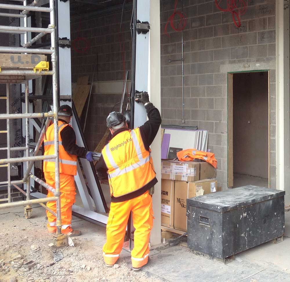 Two workers in orange safety gear guide a large panel at a construction site entrance