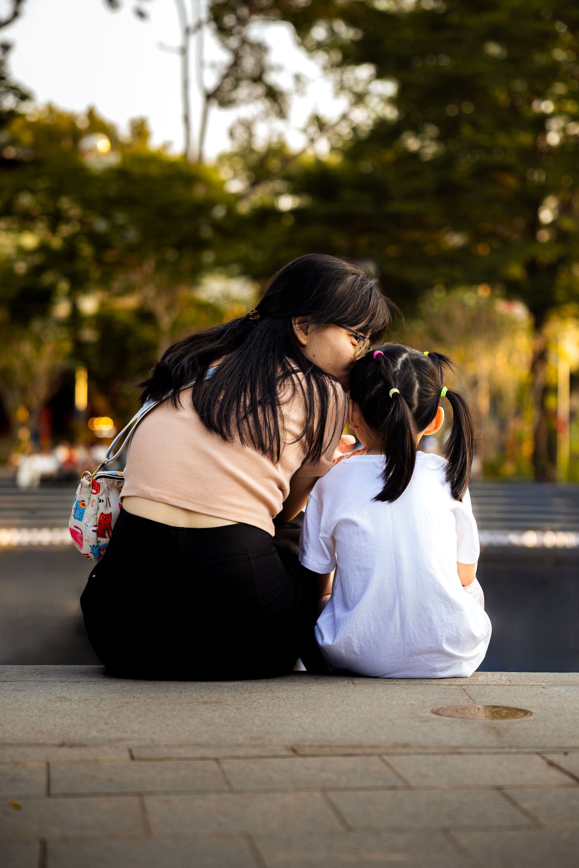 Two people sitting on a sidewalk bench, leaning toward each other in a tree-lined park.