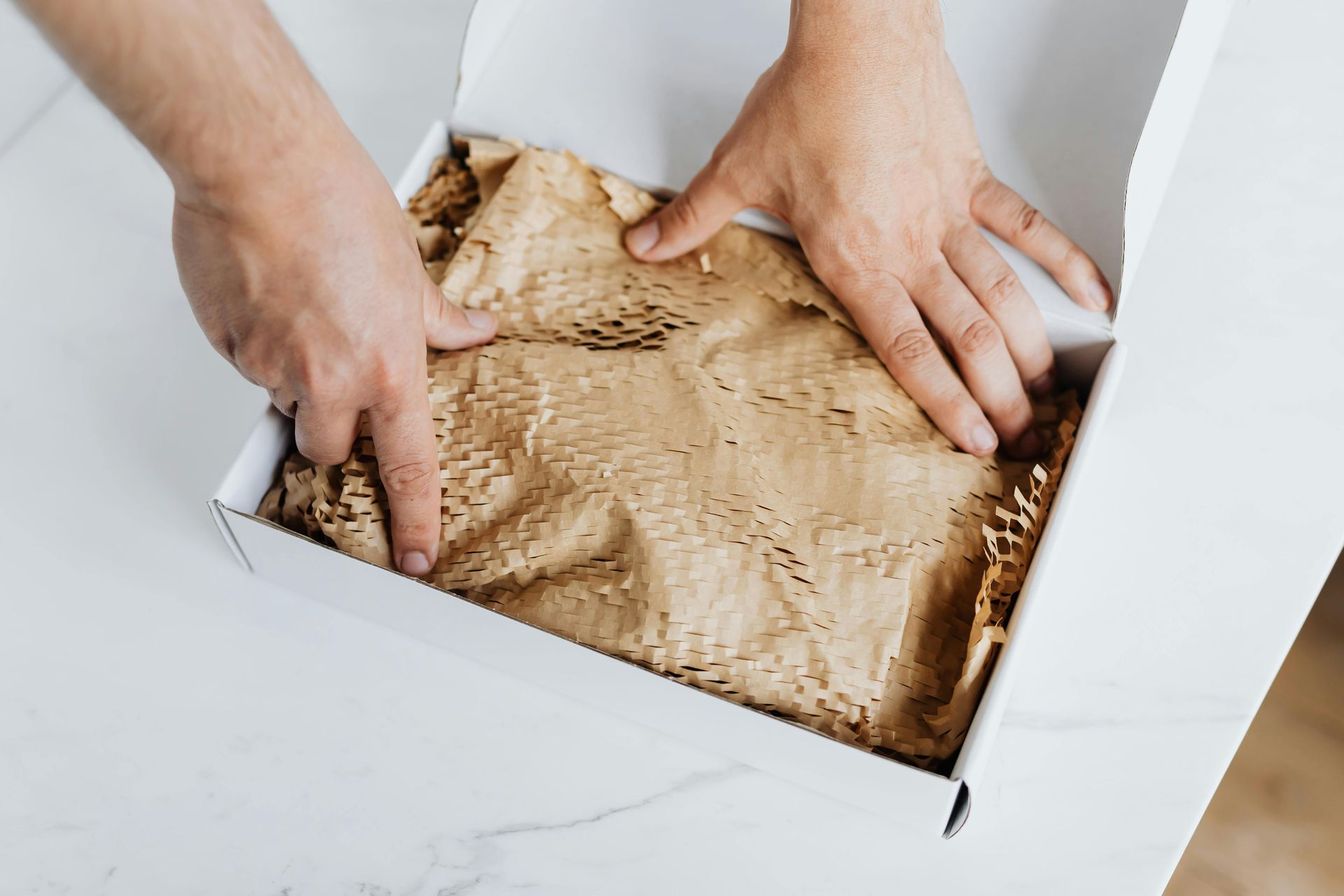 Hands pressing down on brown honeycomb packaging paper inside a white cardboard box on a white surface.