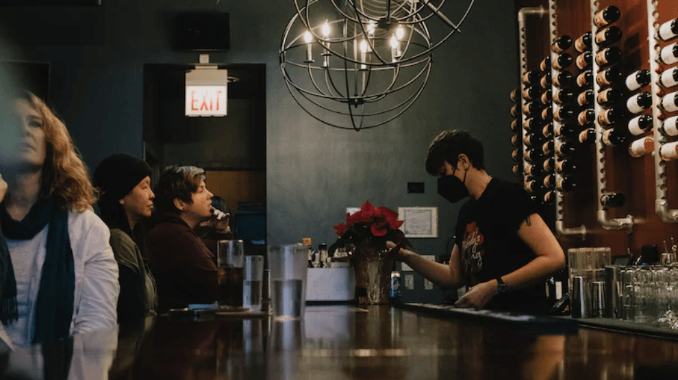 A woman wearing a mask is serving a drink to a group of people at a bar.