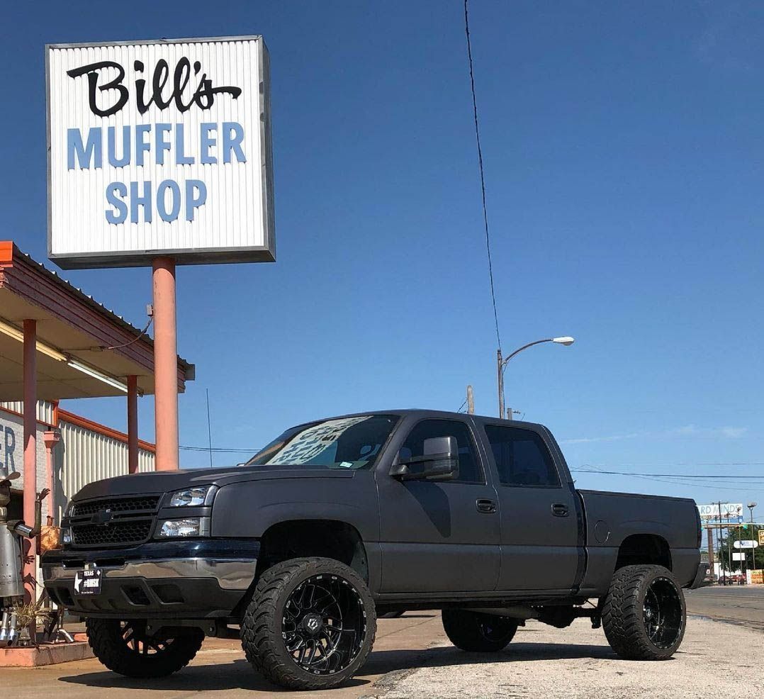 Black lifted pickup truck parked outside Bill's Muffler Shop under a clear blue sky.