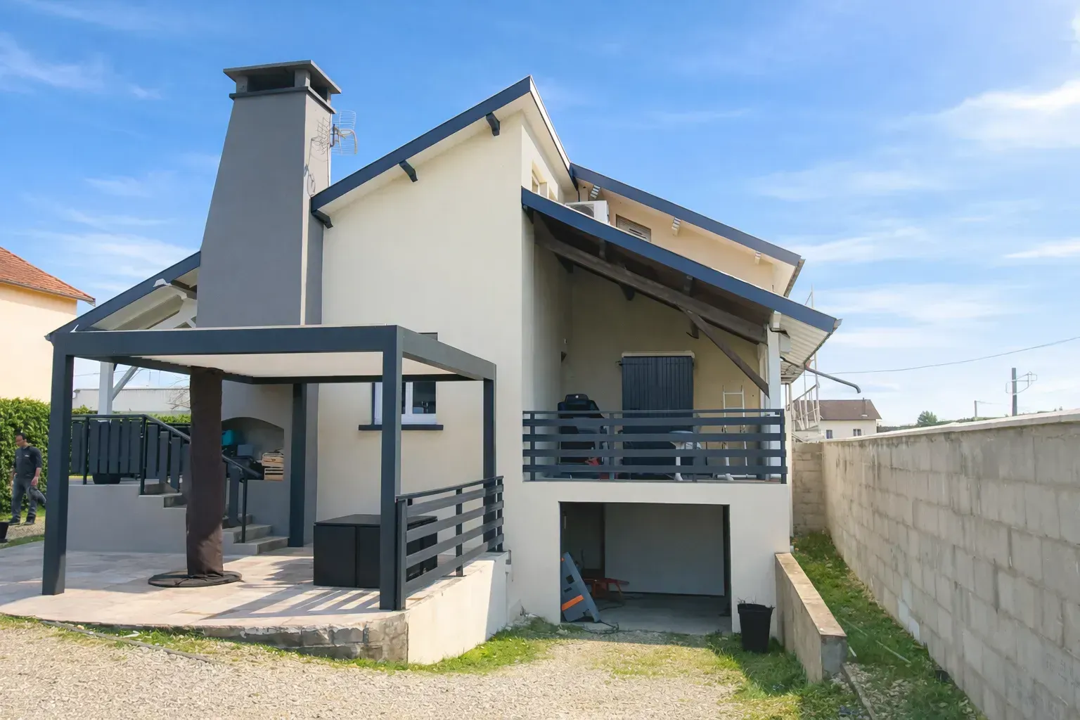 Une maison moderne couleur crème avec une cheminée gris foncé, un patio couvert, une terrasse surélevée et un garage de plain-pied.