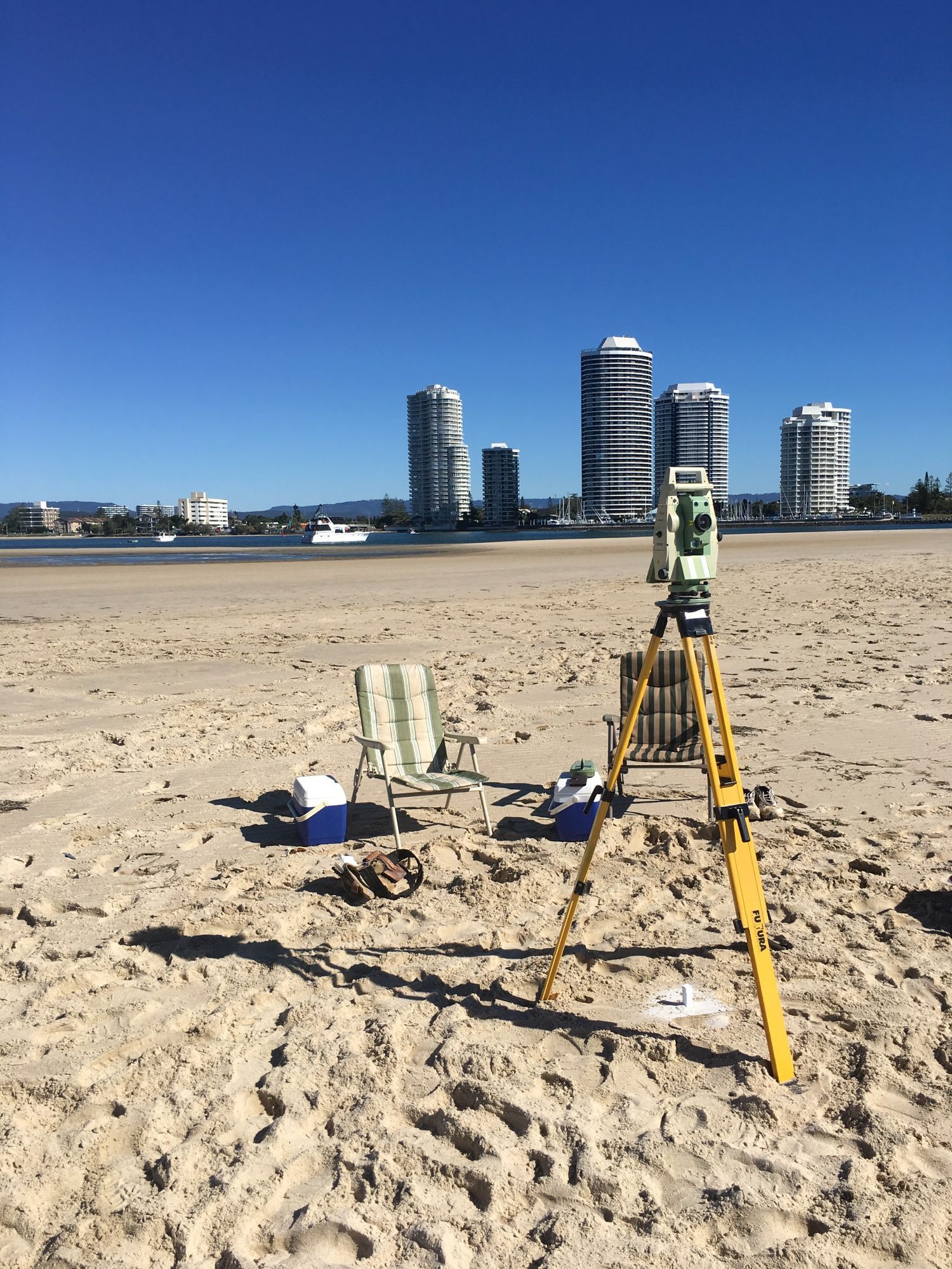 Survey equipment on a sandy beach with tall buildings in the background under a blue sky.