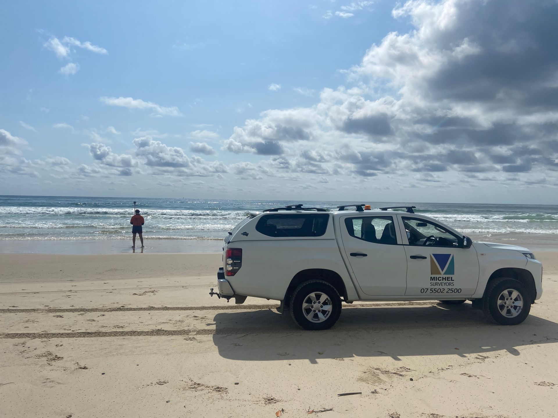 White truck parked on a sandy beach; person wading in the ocean under a partly cloudy sky.