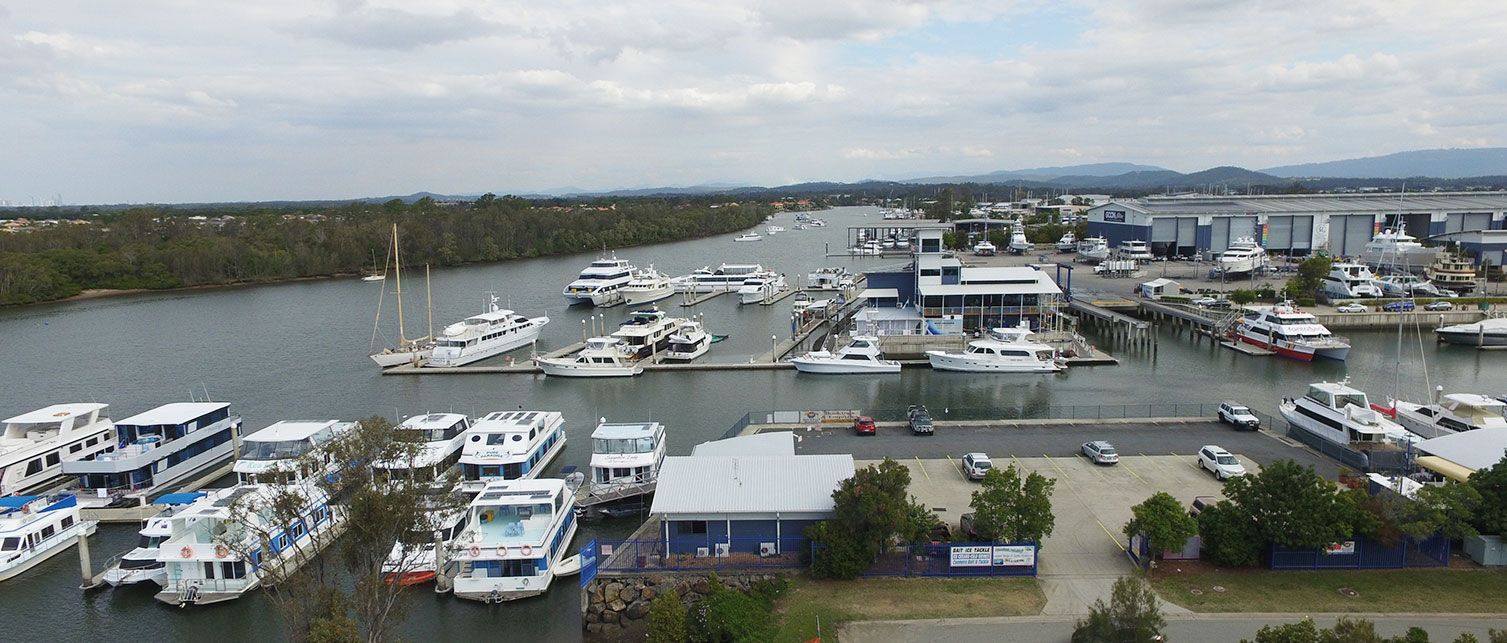 Boats docked at a marina with buildings and a river in the background. Overcast sky.