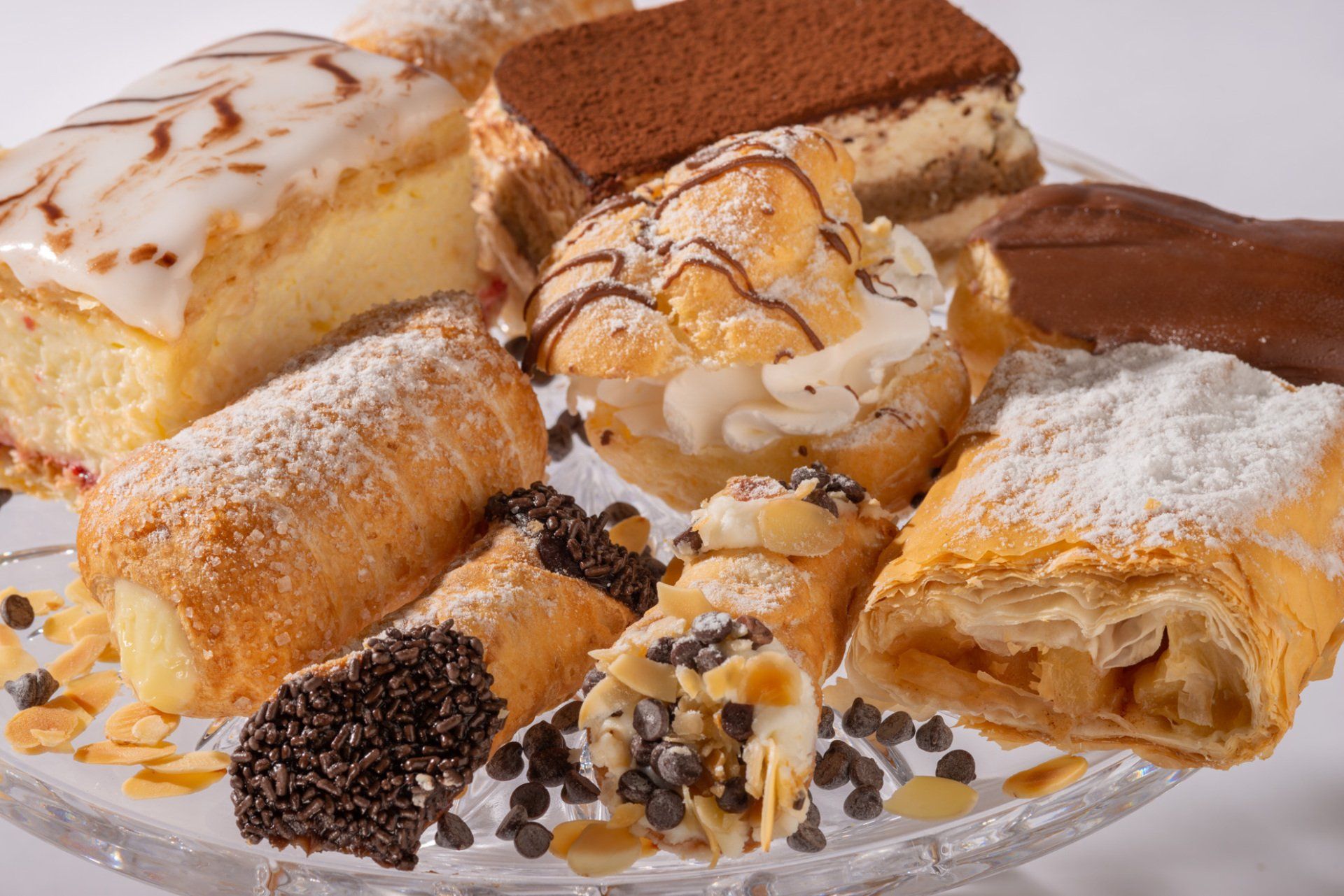 Assorted pastries and cream-filled desserts on a glass plate, dusted with powdered sugar and chocolate drizzle