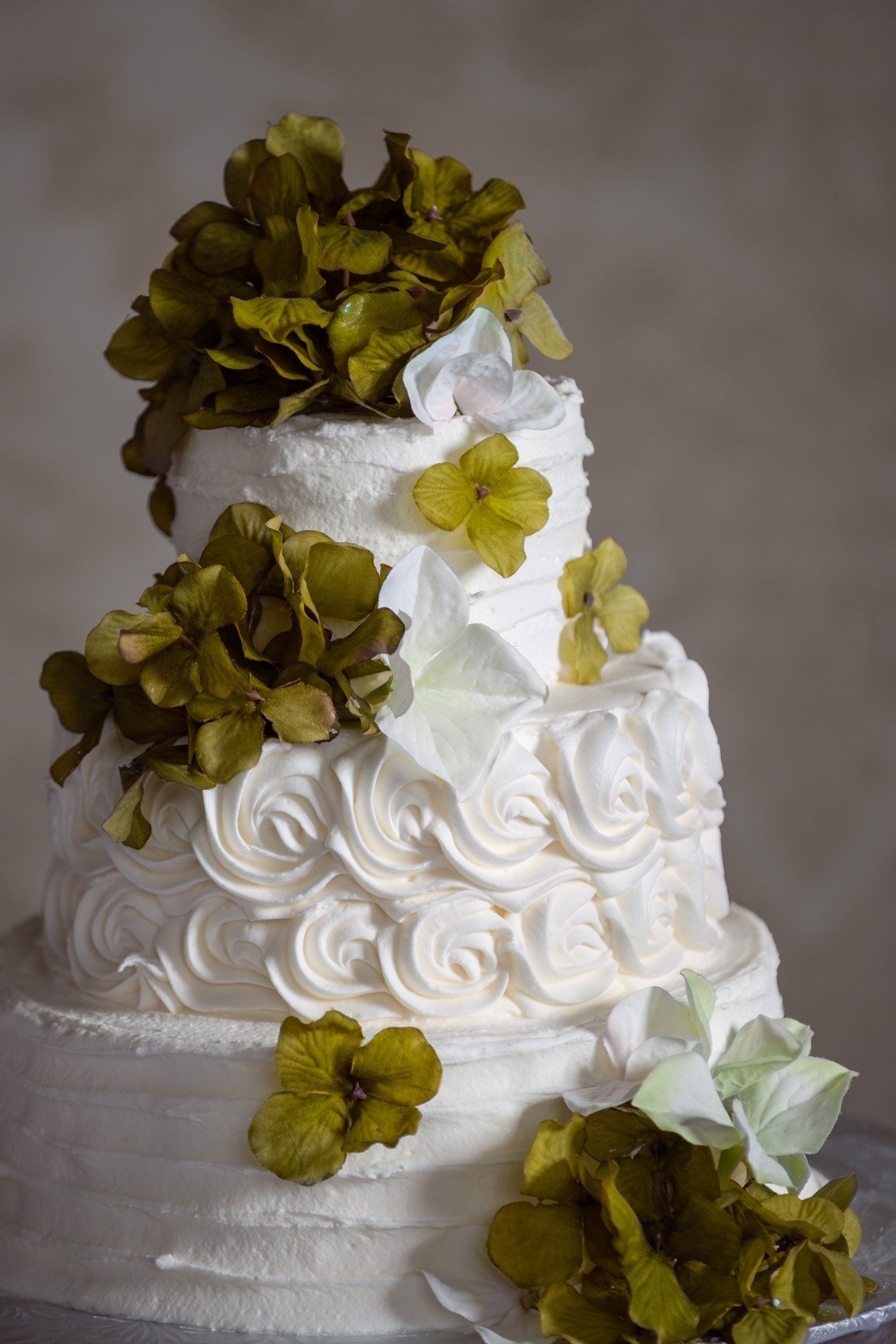 White tiered cake with white frosting and olive-green flowers on a neutral background