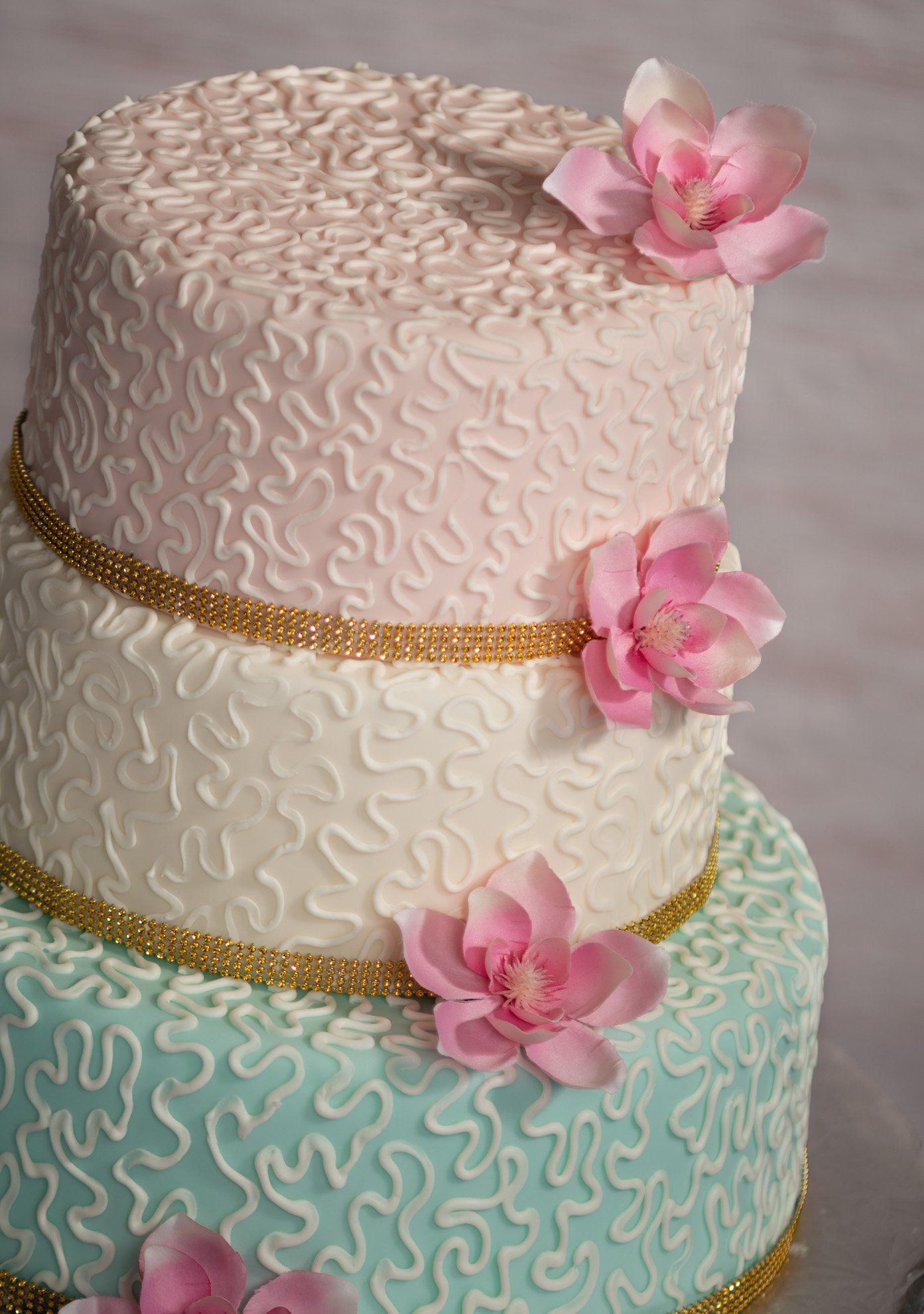 Three-tier cake with white and teal icing, gold trim, and pink flowers on top and sides