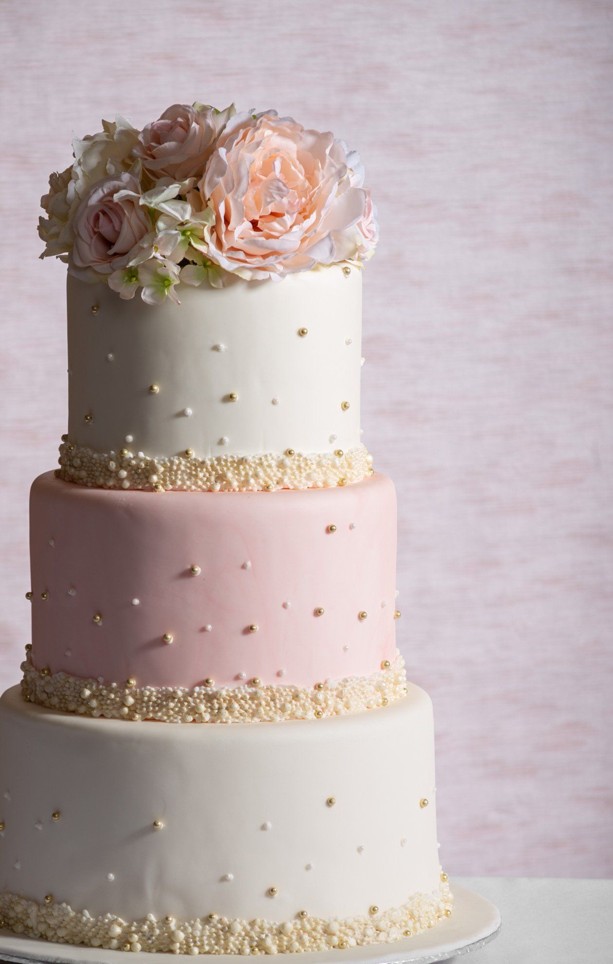 Three-tier pastel wedding cake with flowers on top and gold accents on a pink background