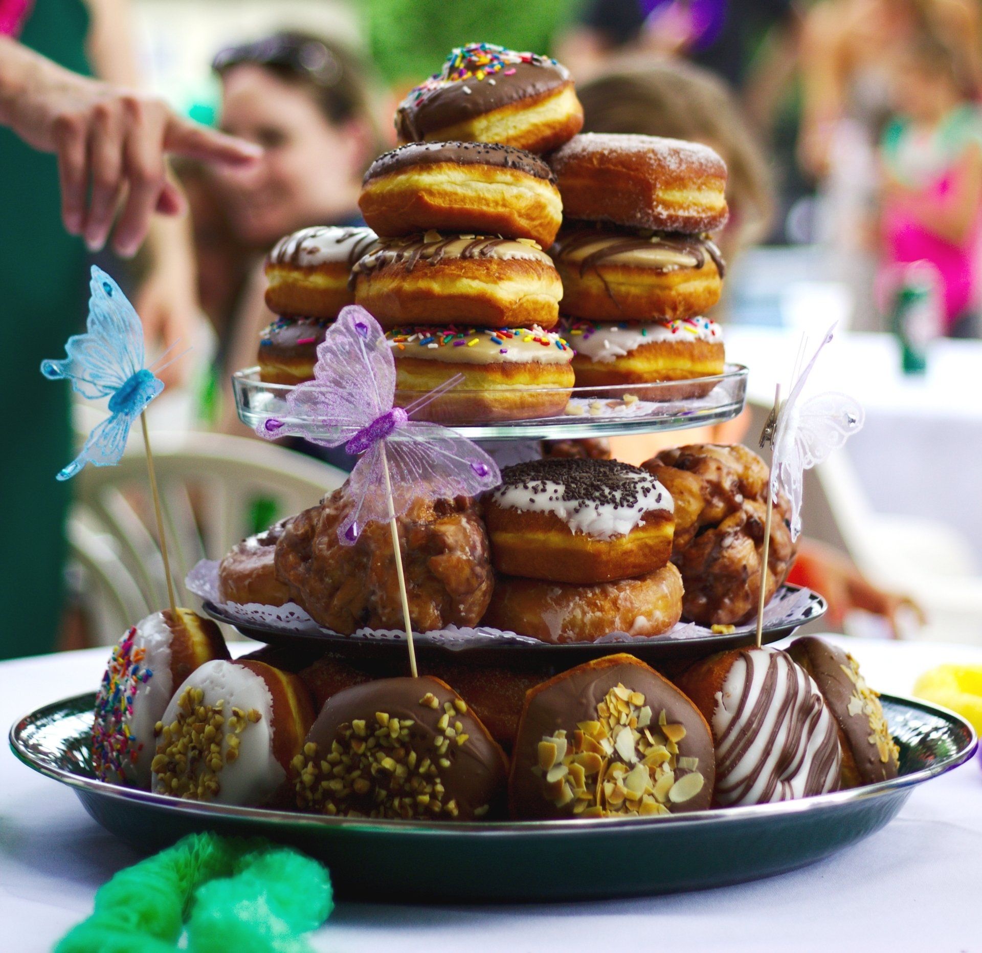 Tiered tray of assorted decorated donuts at an outdoor dessert table, with a hand reaching in from the left.