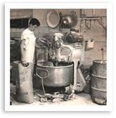 Two workers operate a large industrial mixer in a factory, with metal barrels nearby.