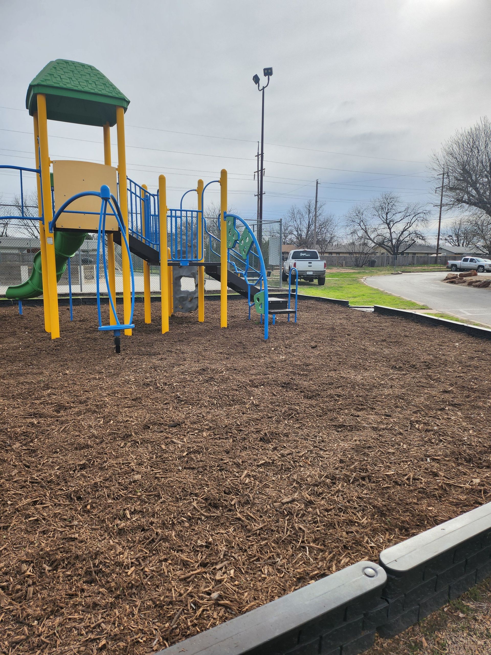 Playground with blue, yellow, and green structure on wood chips, under a cloudy sky.
