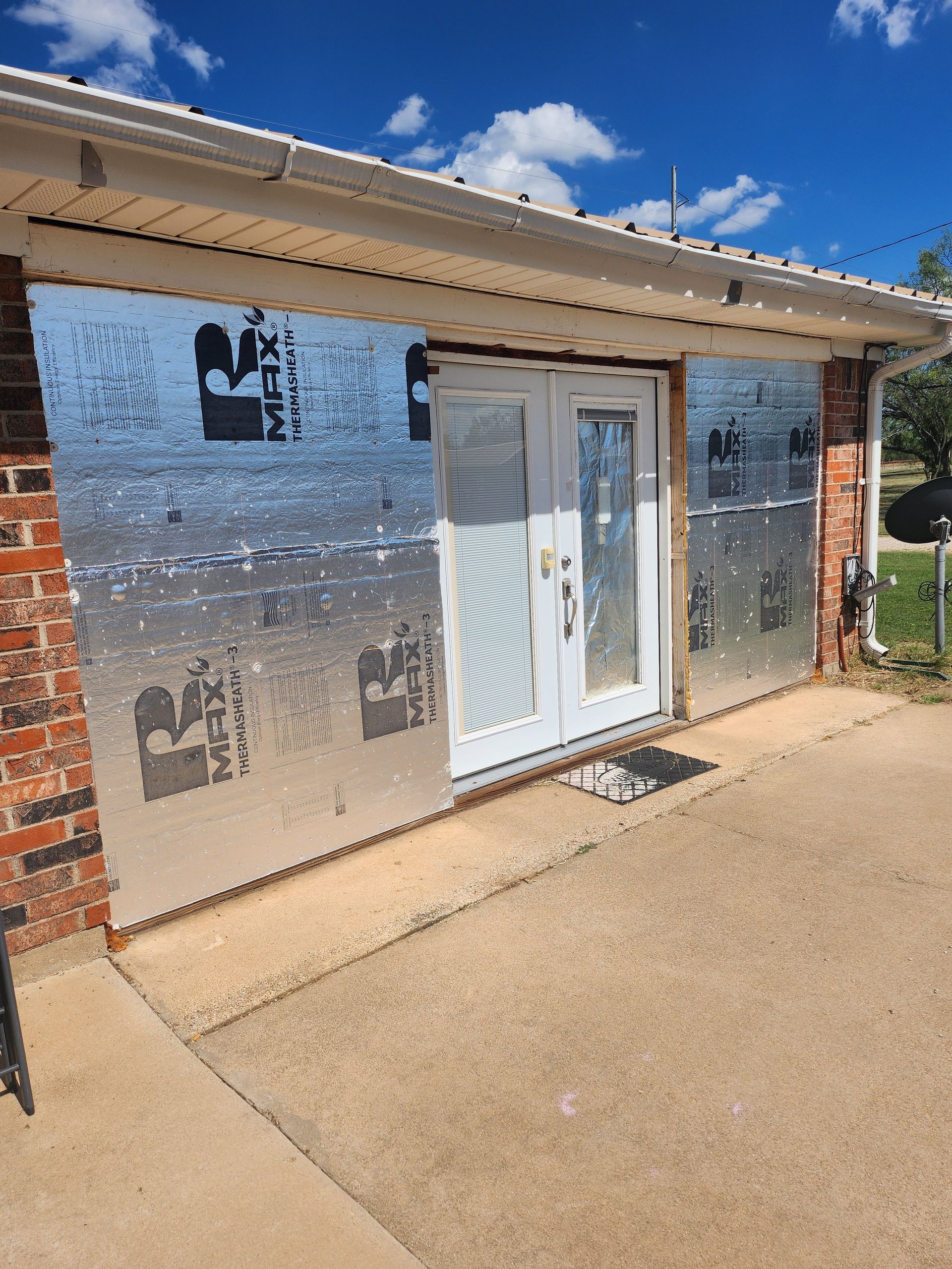 Exterior view of a brick building with a set of white French doors. Silver insulation covers the wall around the doors.