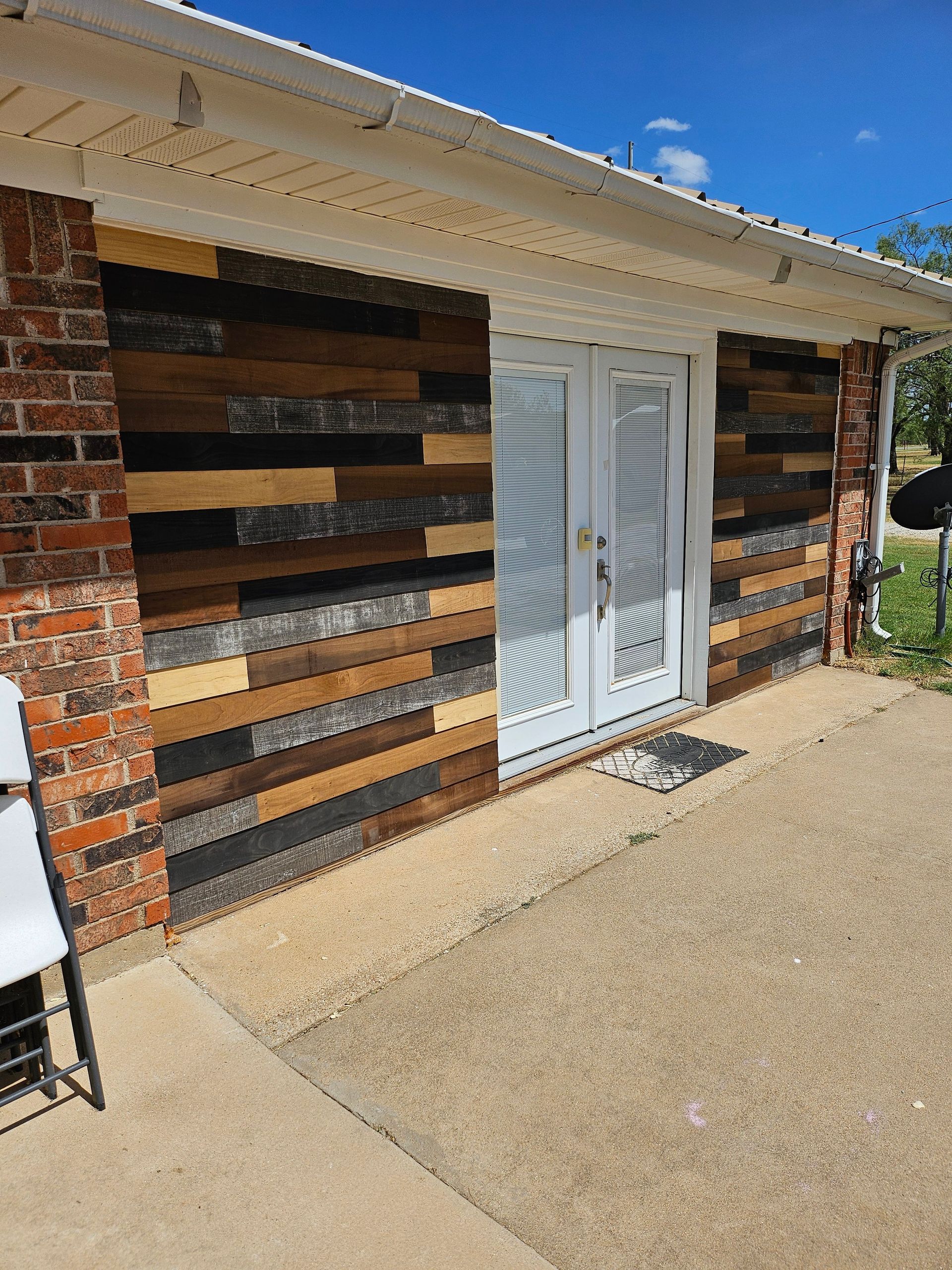 Exterior wall with mixed wood paneling, white door, brick and concrete. Bright day.