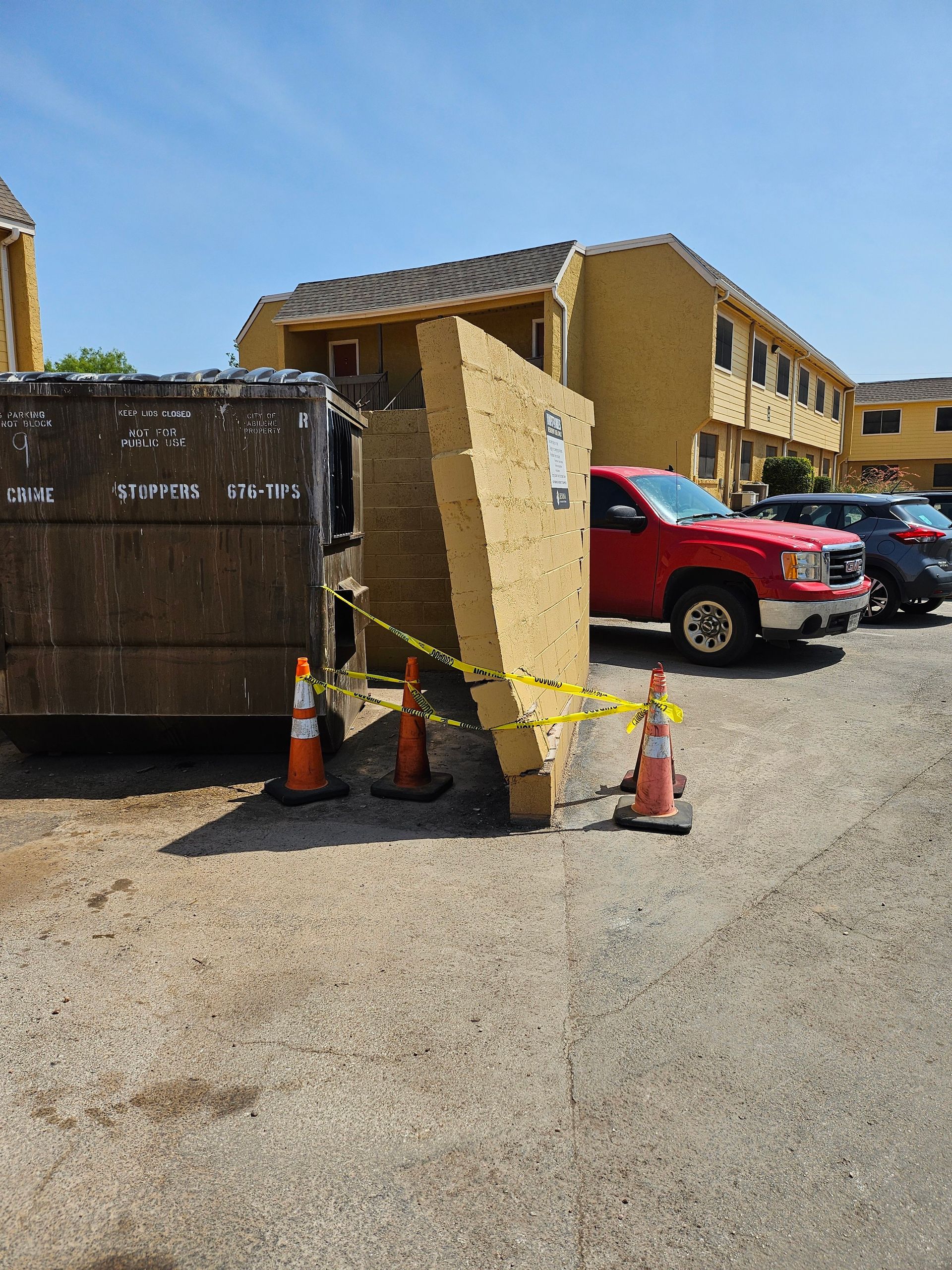 Dumpster with leaning wood panel, cones, and caution tape in a parking lot, with a red truck and apartment buildings.